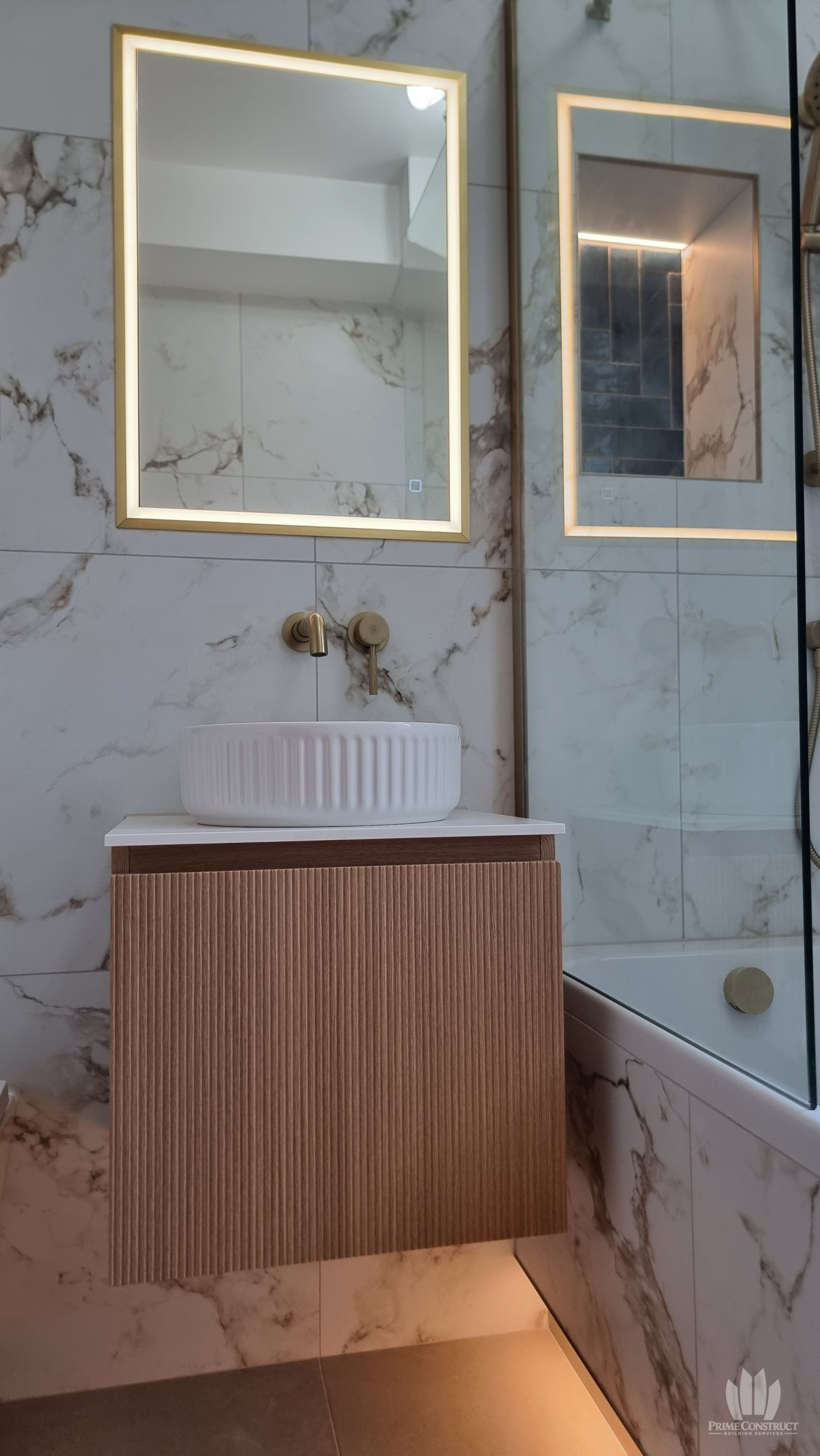 Portrait of fluted wood-tone floating vanity, white fluted basin, gold-tone fittings and illuminated mirror.