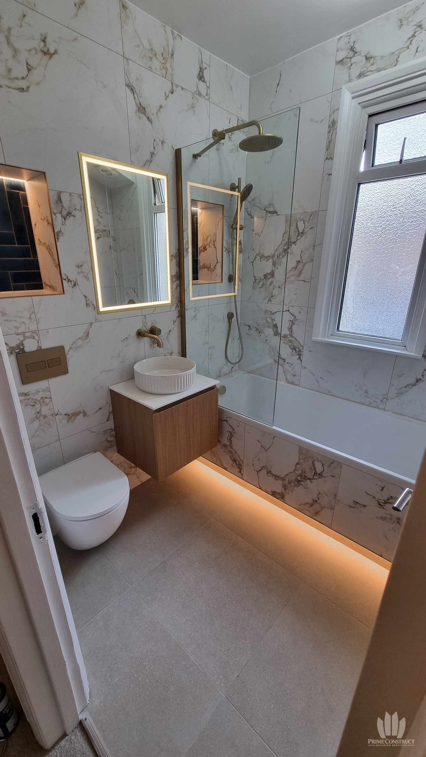 Doorway portrait showing wall-hung WC, fluted wood-tone vanity and basin, bath beneath the window, glass screen and warm under-bath lighting.