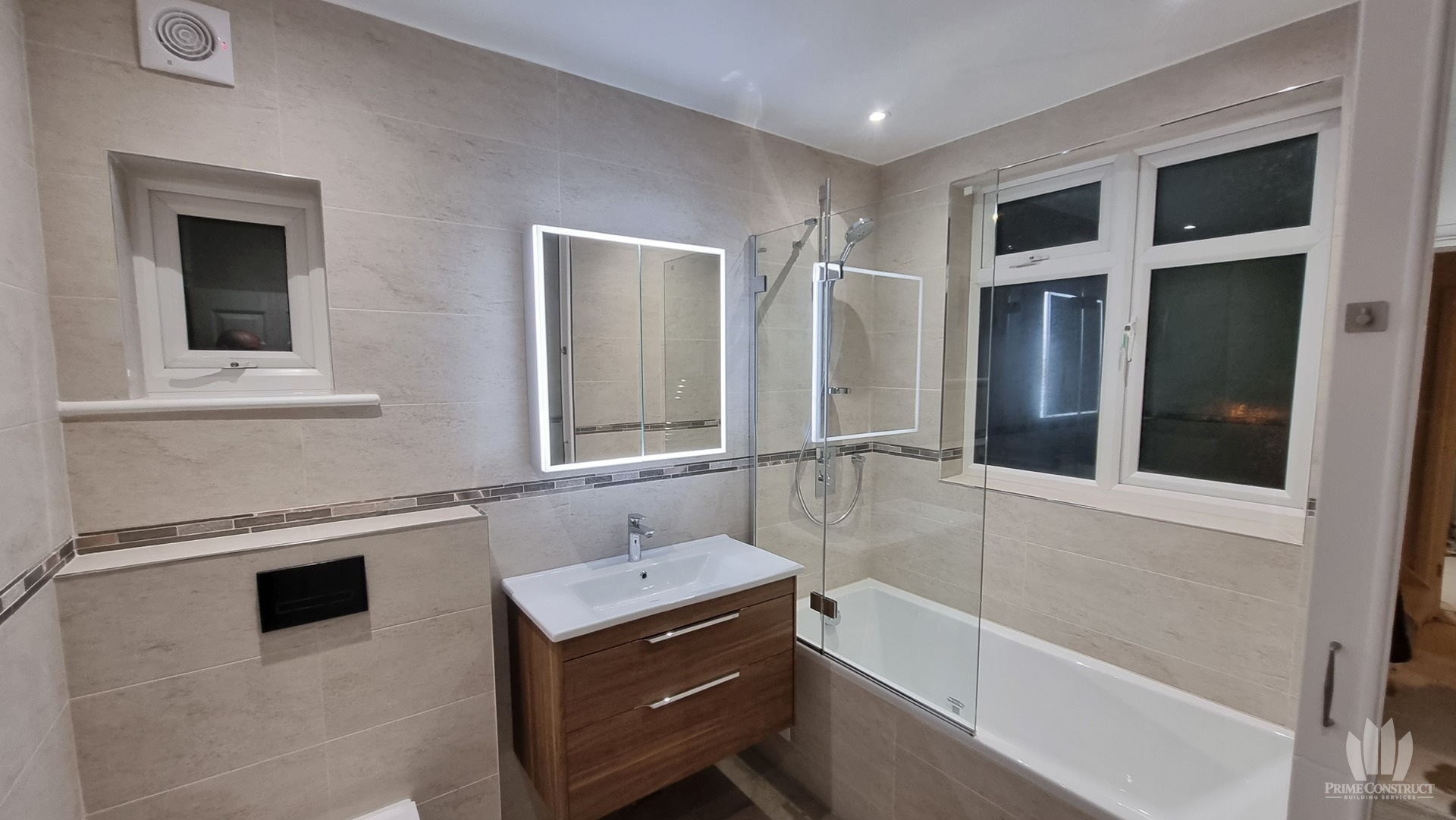 Finished beige-tiled bathroom, bath, wood-tone vanity, concealed WC and mirror.